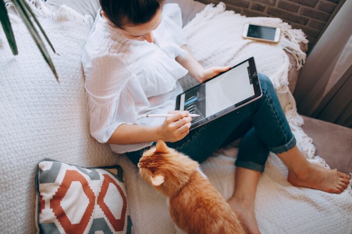 Photo by Helena Lopes woman in white long sleeve shirt and blue denim shorts sitting on bed using tablet computer