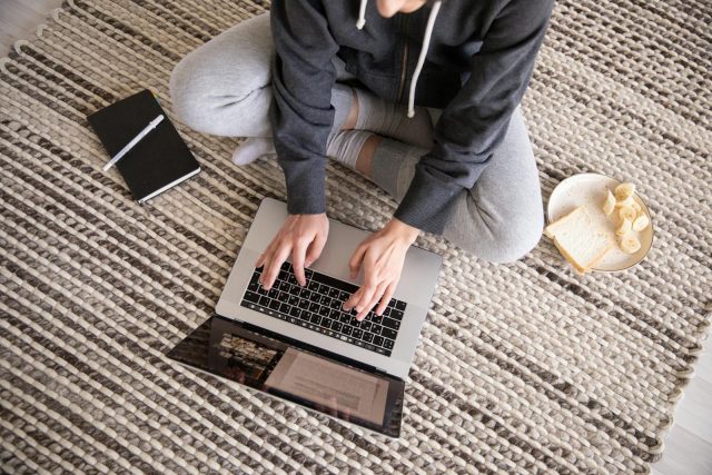 Photo by KATRIN BOLOVTSOVA Woman in casual wear working remotely on a laptop, with a notebook and snack nearby.